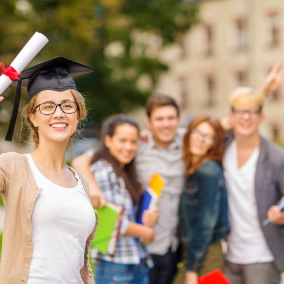 education, campus and teenage concept - smiling teenage girl in corner-cap and eyeglasses with diploma and classmates on the back